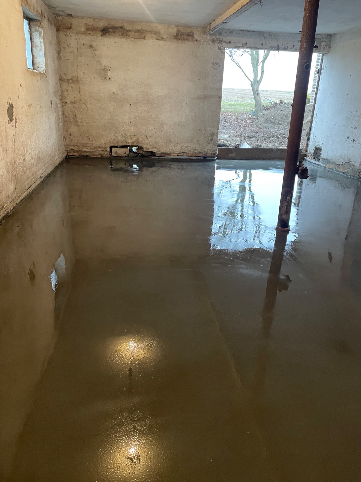 Świeżo wylana, mokra posadzka w surowym pomieszczeniu z widoczną rurą pionową i oknem z widokiem na zewnątrz. Gładka powierzchnia odbija światło.