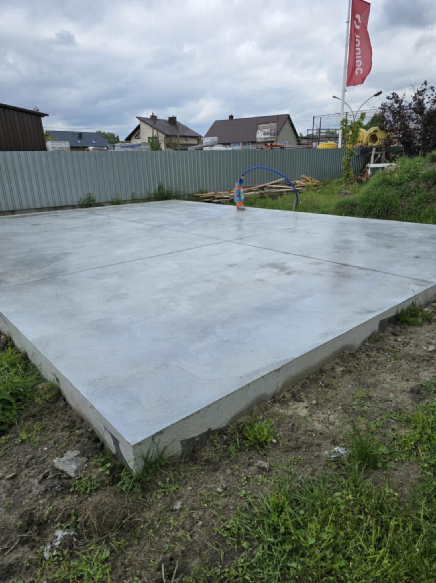 Wylana, szara płyta betonowa na zewnątrz, podzielona na cztery sekcje, z widocznym zbrojeniem i otoczeniem trawiastym. W tle budynki mieszkalne i flaga.