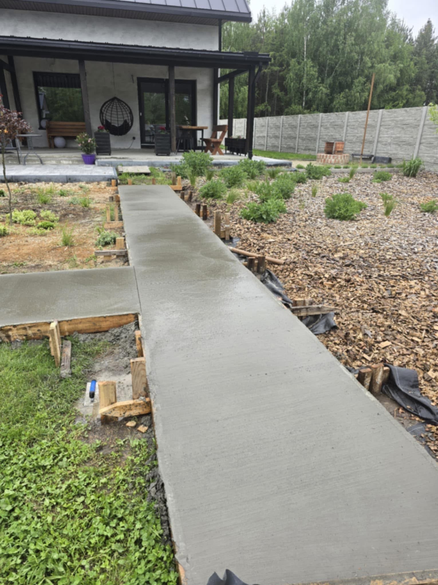 Świeżo wylana, szara ścieżka betonowa z drewnianym szalunkiem prowadząca do domu z tarasem i ogrodem. Widoczne rośliny i kora na ziemi.