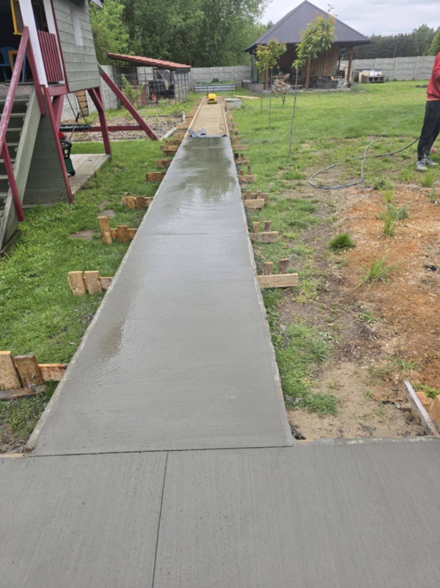 Świeżo wylana, długa, betonowa ścieżka w ogrodzie, z drewnianym szalunkiem po bokach. Widoczny fragment placu zabaw i budynek gospodarczy w tle.