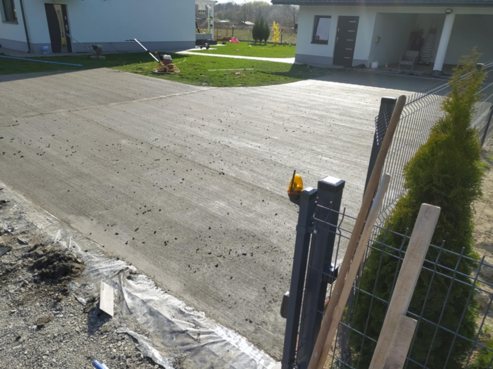 Świeżo wylana, szara posadzka na podjeździe domu jednorodzinnego, z widocznymi śladami po zacieraniu mechanicznym. W tle budynki i zieleń trawnika.