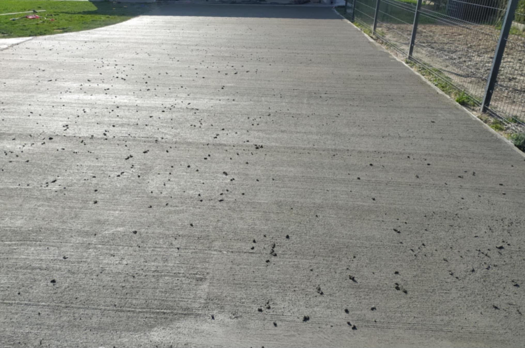 Świeżo wylana, szara posadzka z widocznymi śladami po zacieraniu, z drobnymi zanieczyszczeniami na powierzchni. W tle zielony trawnik i metalowe ogrodzenie.