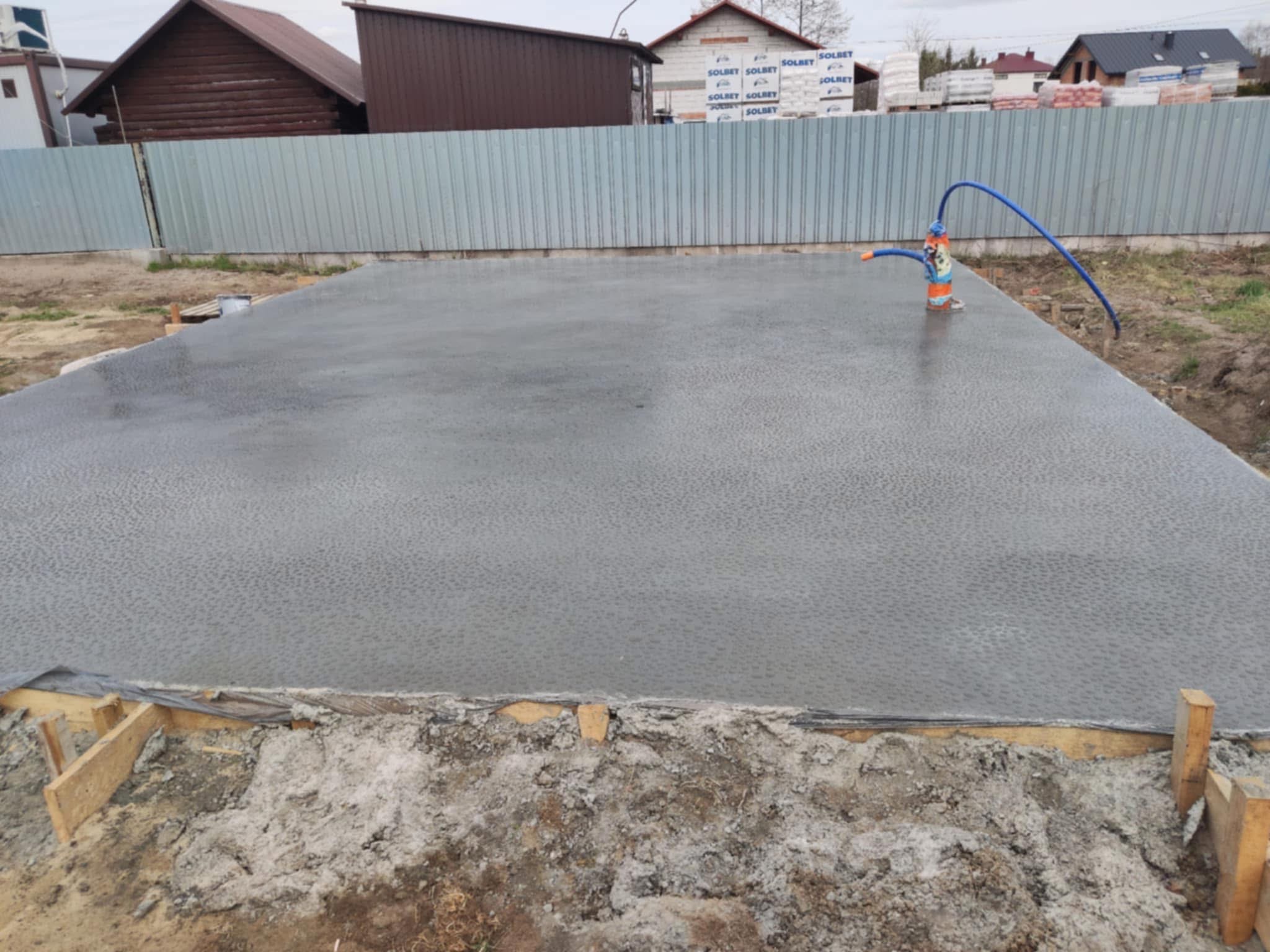 Świeżo wylana, gładka posadzka betonowa na budowie, z widocznym poziomowaniem i niebieskim wężem. W tle ogrodzenie z blachy falistej i budynki.