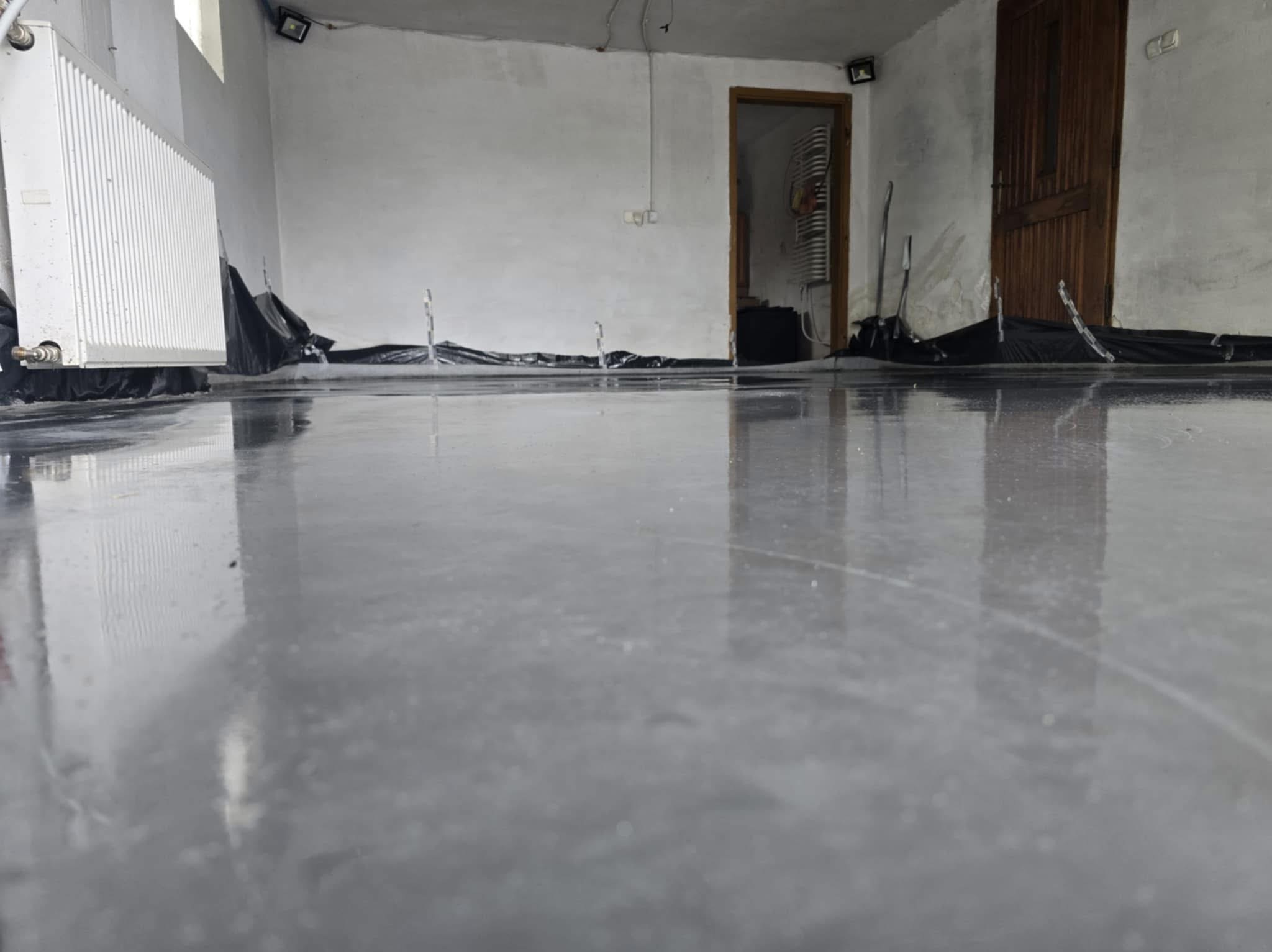 Błyszcząca, szara posadzka w surowym wnętrzu z grzejnikiem i otwartymi drzwiami. Folia ochronna przy ścianach. Świeżo wylana, gładka powierzchnia.