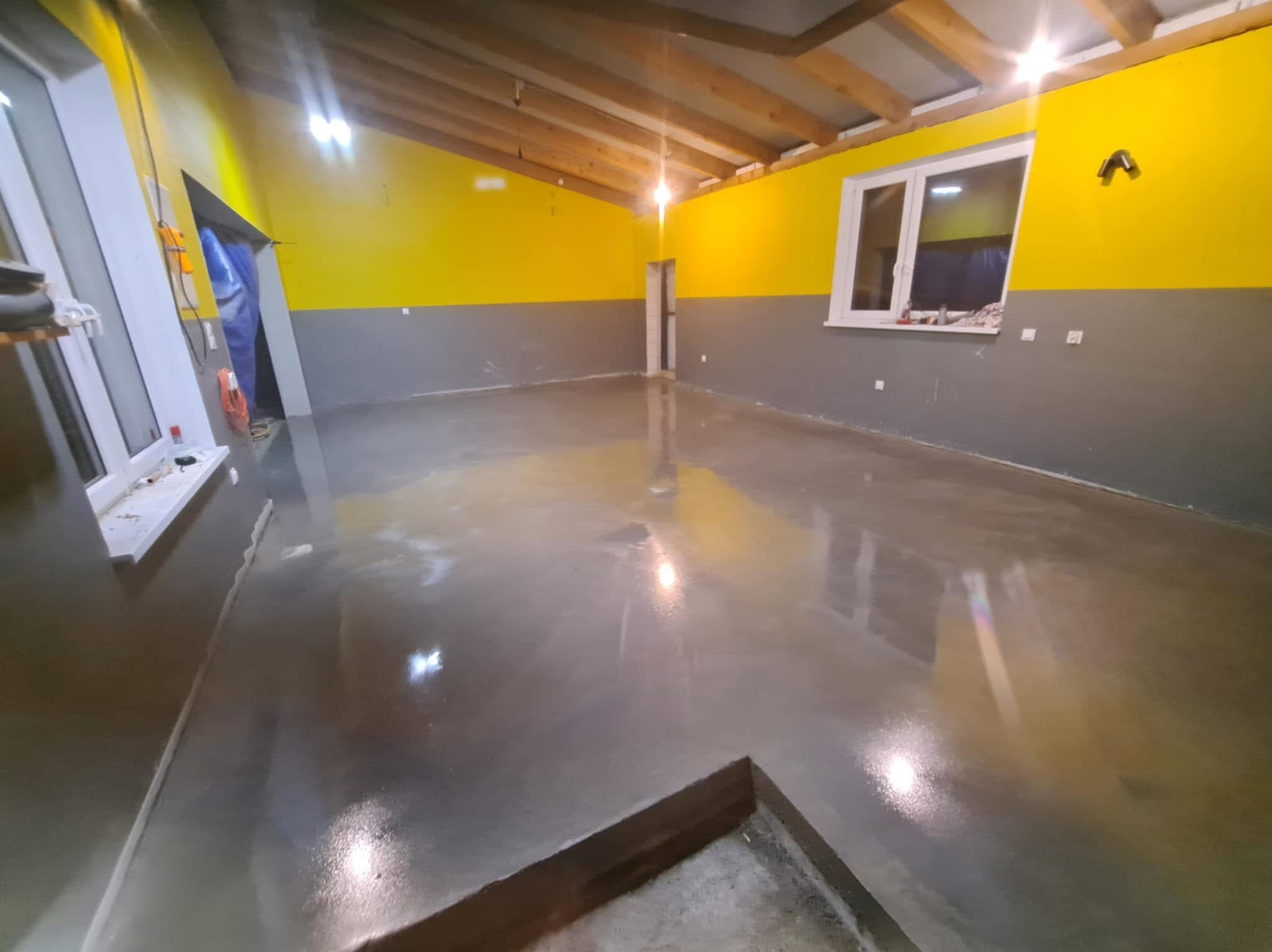 Błyszcząca, szara posadzka w nowo wykończonym pomieszczeniu z żółto-szarymi ścianami i drewnianym stropem. Widoczne okno i drzwi.