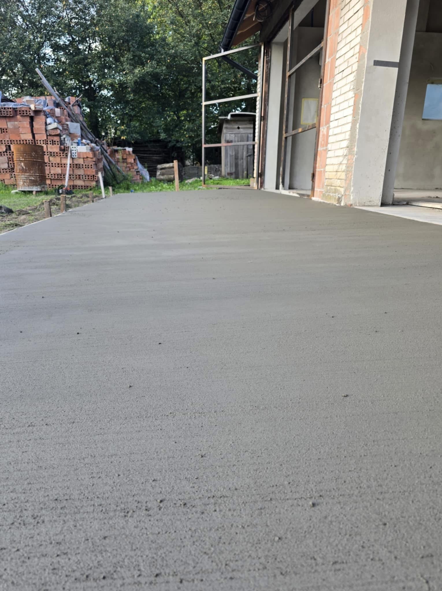 Świeżo wylana, gładka posadzka przemysłowa w surowym stanie, z widocznymi śladami po narzędziach, cegły w tle, częściowo otwarty budynek w tle.