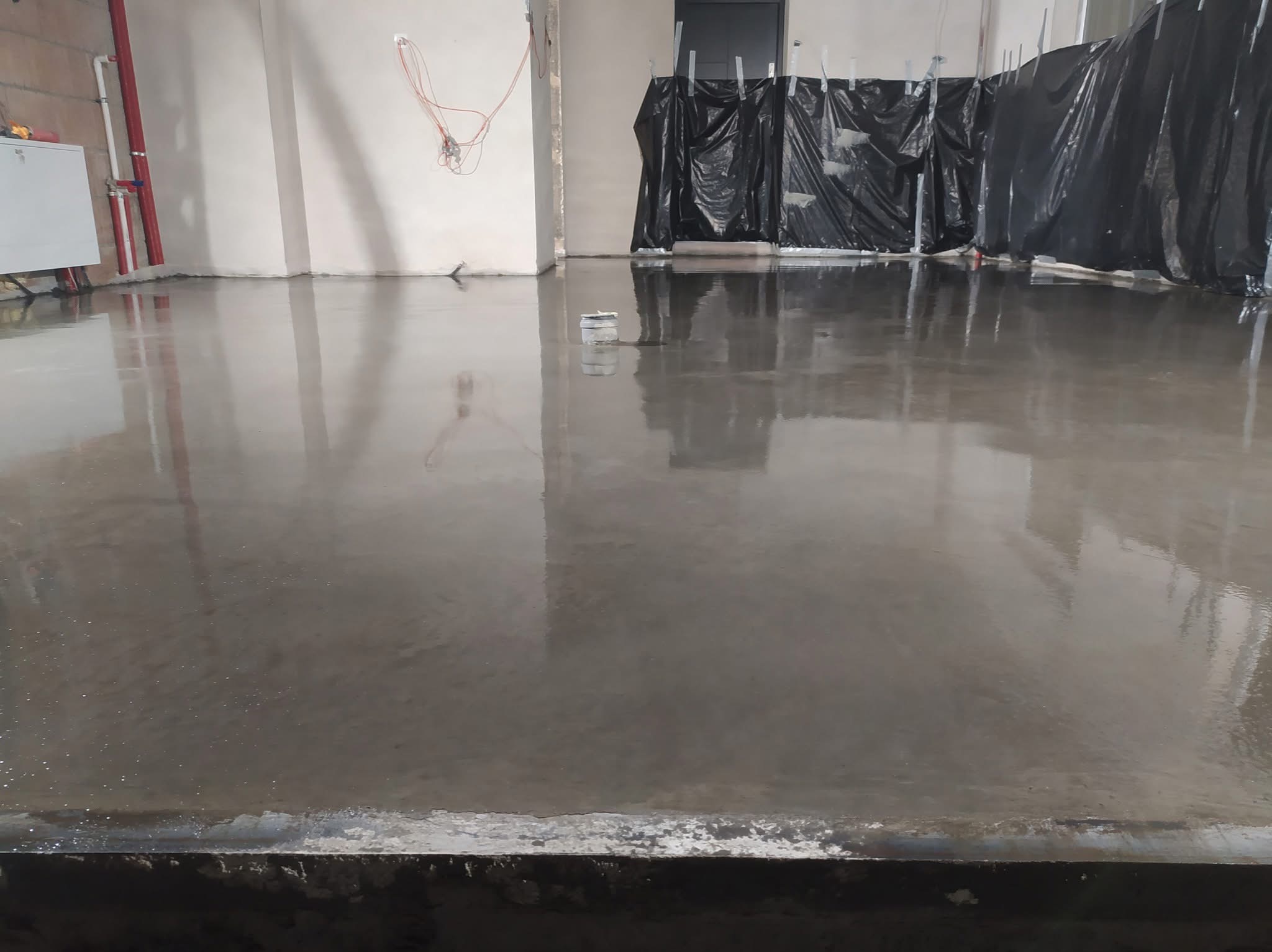 Świeżo wylana, gładka posadzka przemysłowa w surowym wnętrzu. Widoczne instalacje na ścianach i zabezpieczenia folią. Błyszcząca powierzchnia odbija światło.