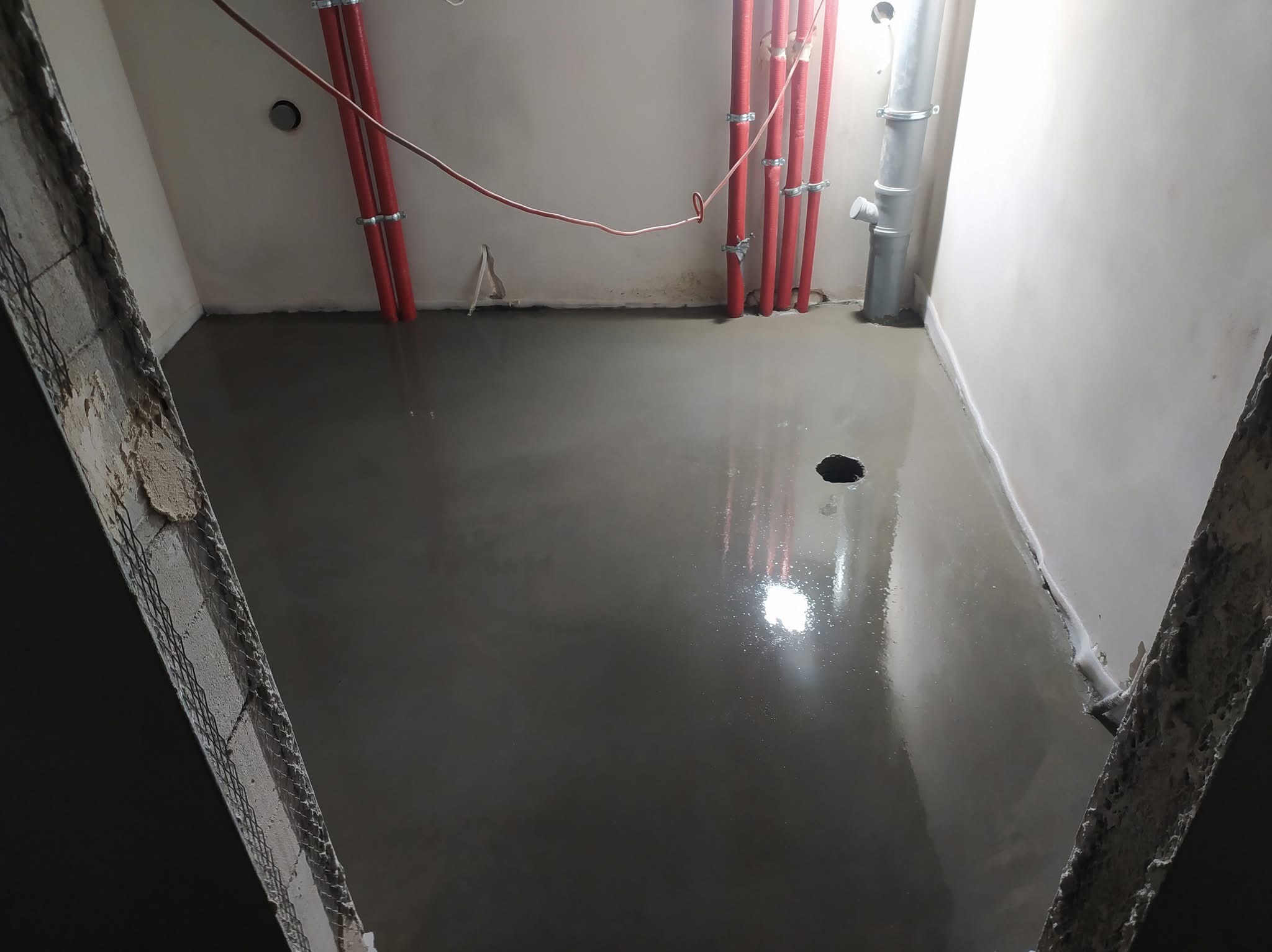Świeżo wylana, gładka, szara wylewka podłogowa w pomieszczeniu z czerwonymi rurami przy ścianie. Widoczne ślady wilgoci i odbicia światła na powierzchni.