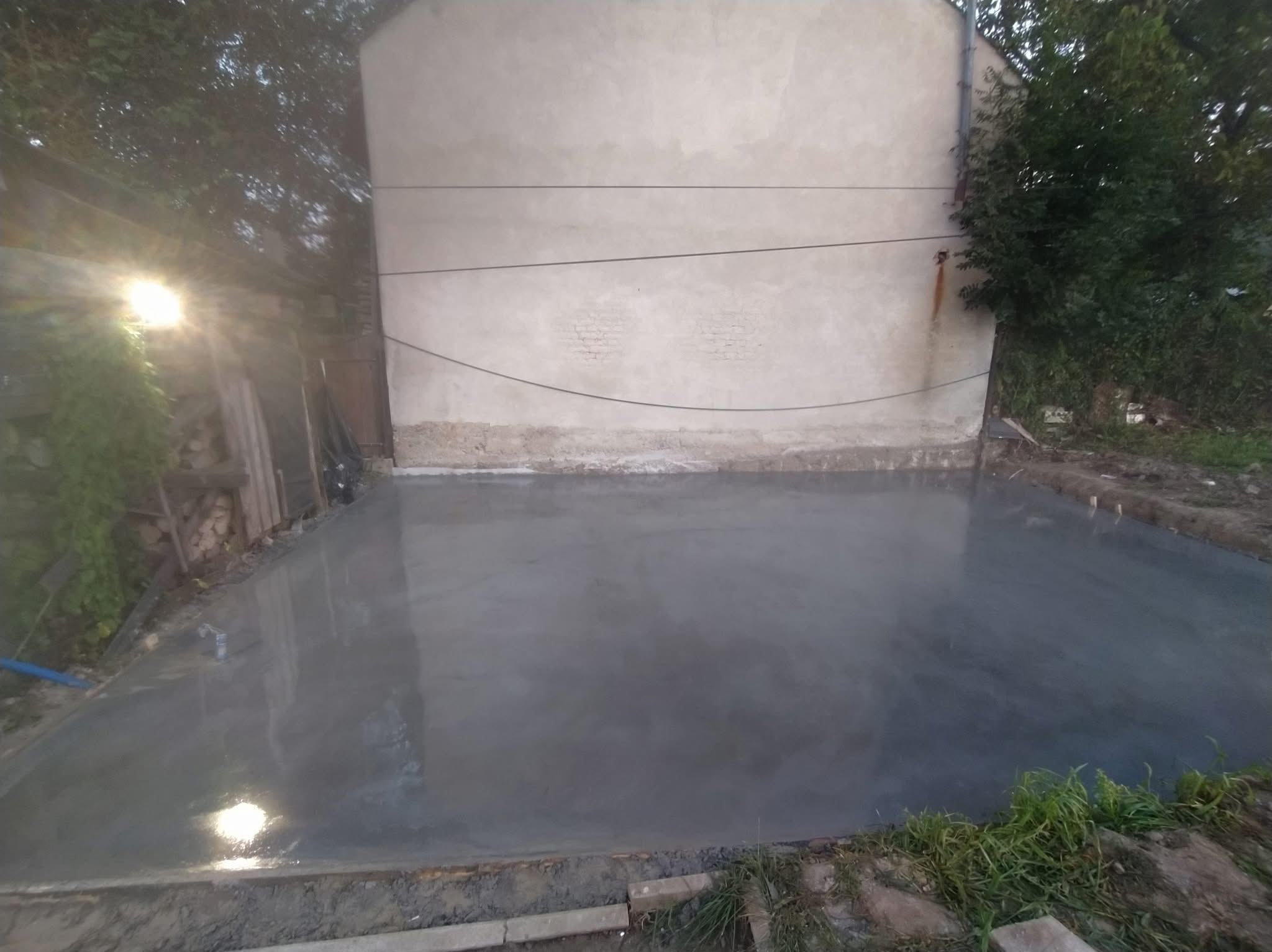 Wylana, świeża, szara posadzka betonowa na zewnątrz, oświetlona lampą budowlaną. Widoczne ślady po narzędziach i obrzeża. W tle ściana budynku i zieleń.