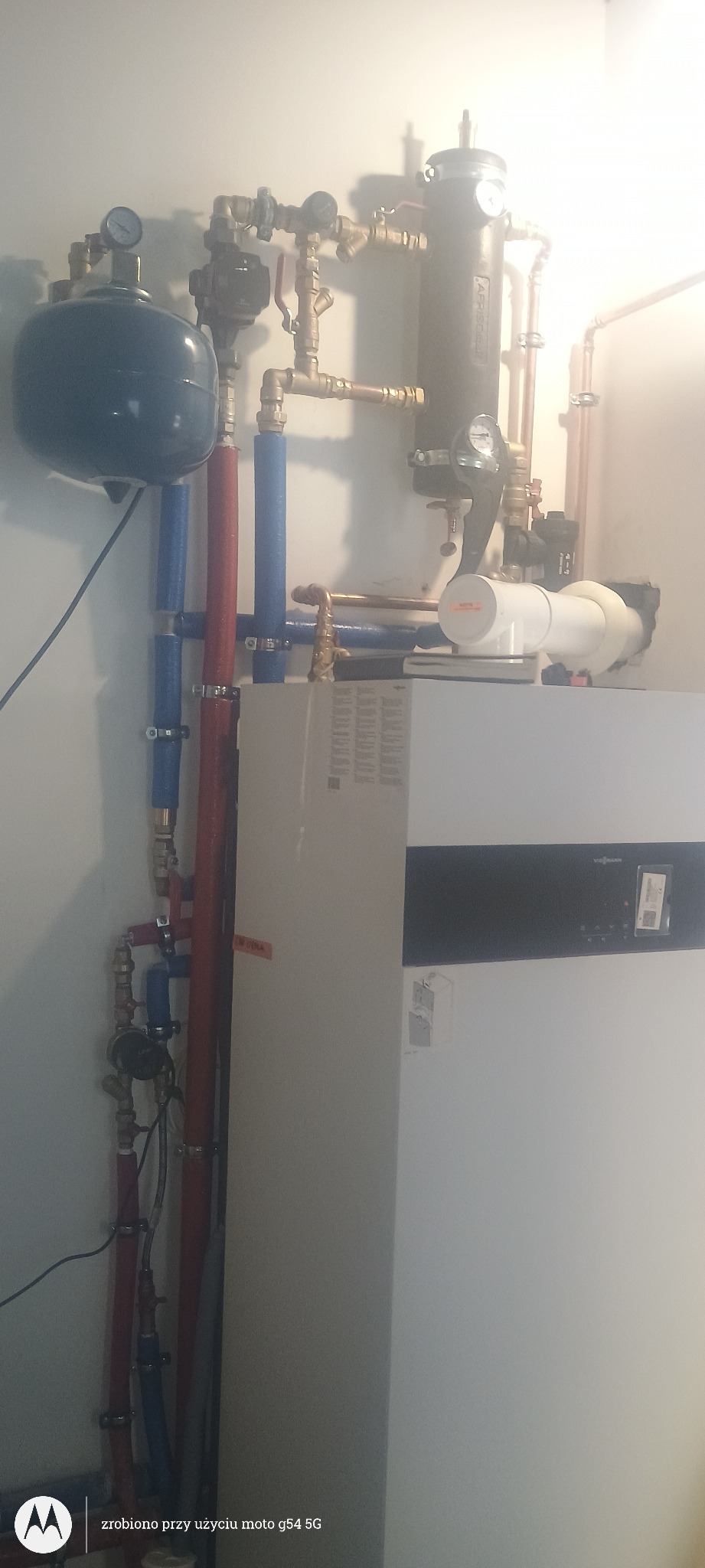 Nowoczesny piec gazowy Viessmann z kompletną instalacją: zbiornik wyrównawczy, pompy obiegowe, rury miedziane i PEX w kolorach niebieskim i czerwonym. Białe ściany w tle.