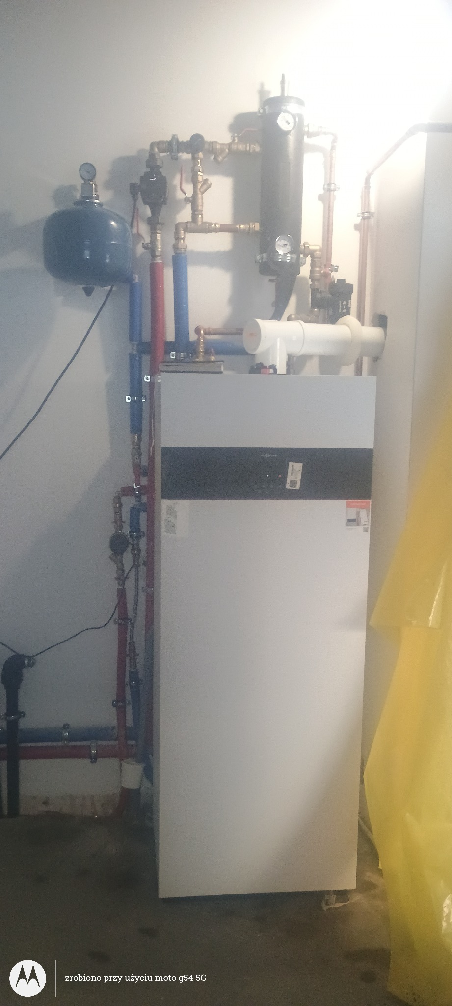 Biały piec grzewczy Viessmann z podłączonymi rurami miedzianymi i elementami instalacji w kotłowni. Widoczne zawory, manometry i zbiornik wyrównawczy.