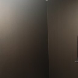 Maksym Nesterenko - Pomieszczenie po malowaniu ścian na ciemny, grafitowy kolor. Widoczny narożnik pokoju z szarą podłogą i fragmentem zabudowy po prawej stronie.