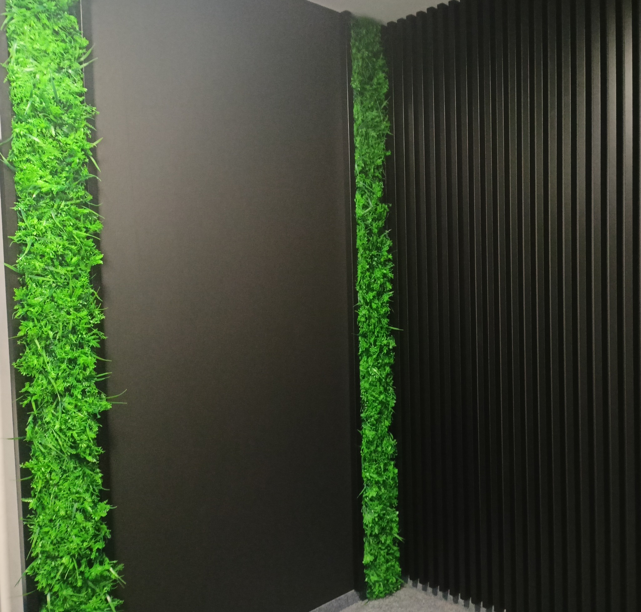 Nowoczesna aranżacja ściany z pionowymi panelami i pasami zieleni w pomieszczeniu. Dekoracja wnętrza z elementami roślinnymi i ciemną kolorystyką.