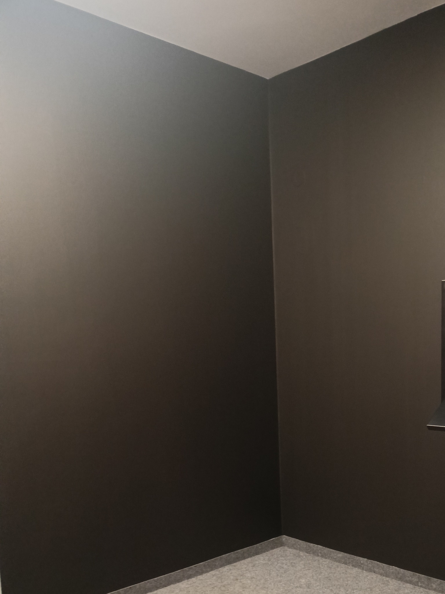 Pomieszczenie po malowaniu ścian na ciemny, grafitowy kolor. Widoczny narożnik pokoju z szarą podłogą i fragmentem zabudowy po prawej stronie.