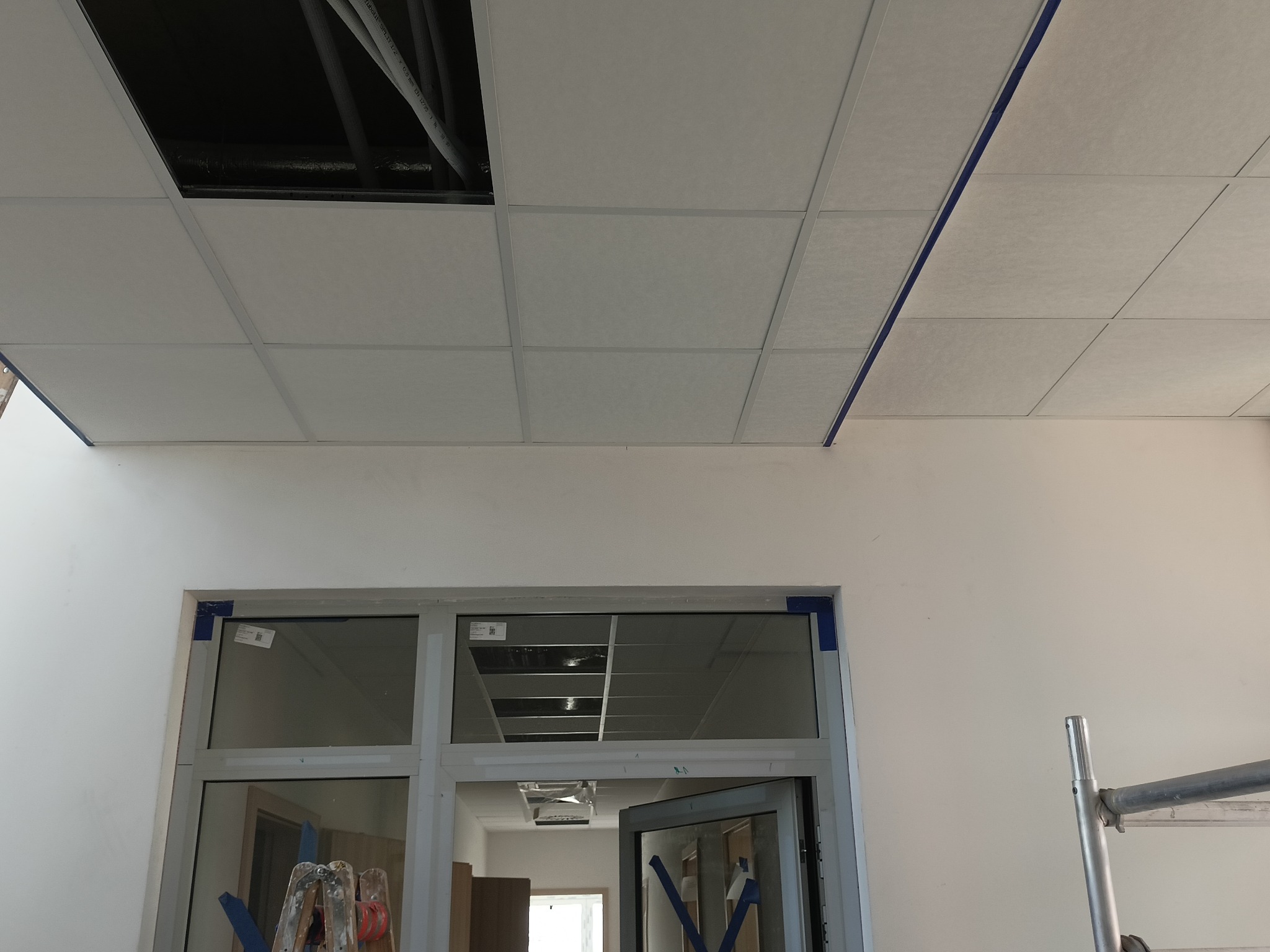 Montaż sufitu podwieszanego kasetonowego w biurze, widoczne kable i rury nad sufitem, okno zabezpieczone taśmą malarską, fragment rusztowania.