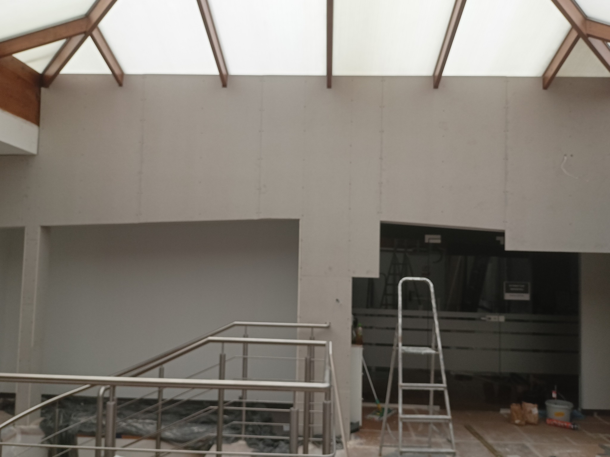 Wnętrze z płytami gipsowo-kartonowymi pod przeszklonym dachem, widoczna drabina i fragment schodów z metalową barierką. Etap prac wykończeniowych.