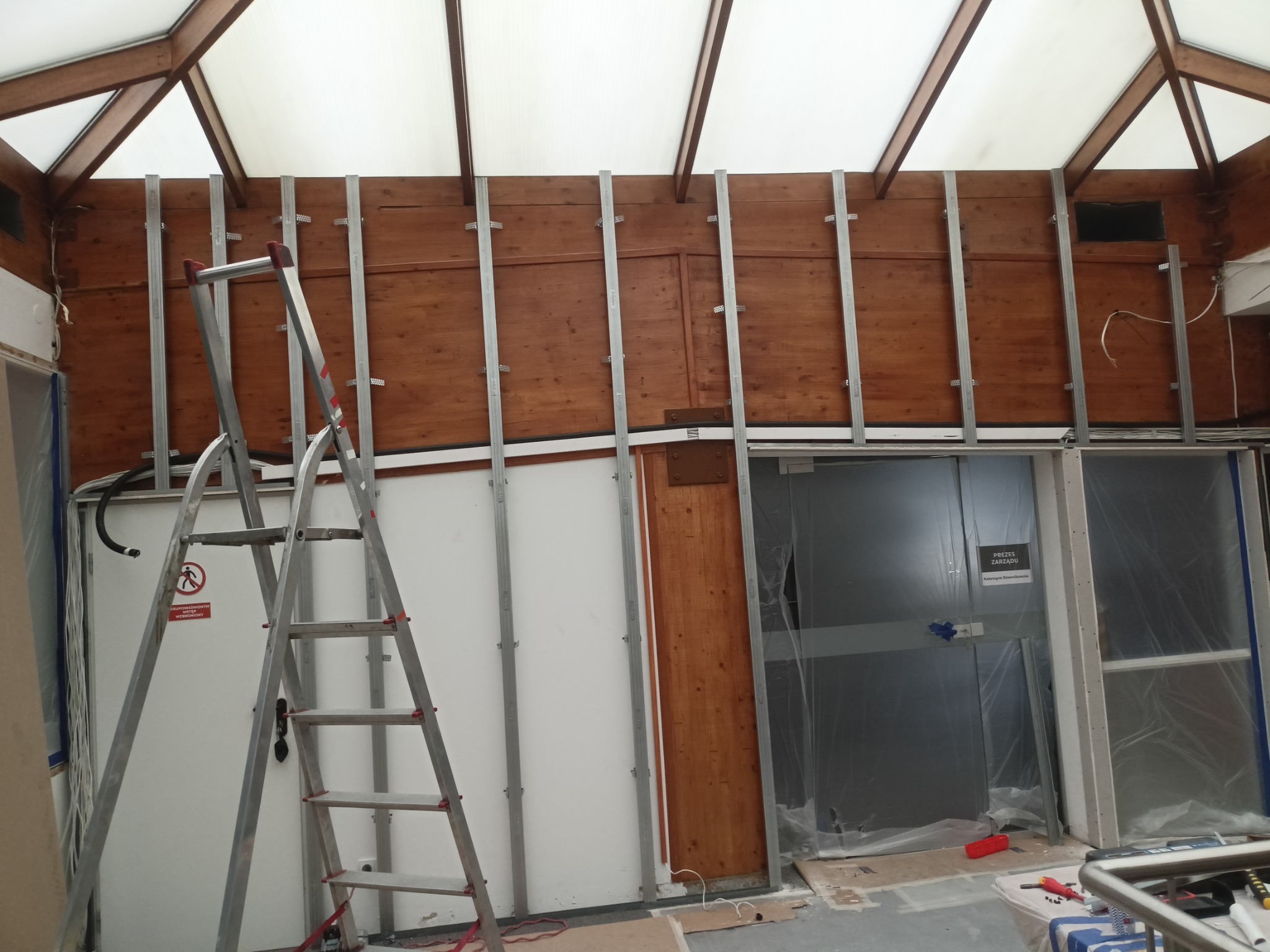 Montaż stelażu pod płytę G-K na drewnianej ścianie w pomieszczeniu ze świetlikiem dachowym. Metalowa drabina oparta o ścianę. Częściowo zamontowana płyta.