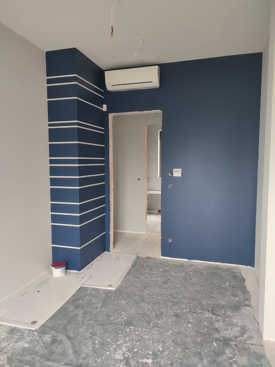 Pomieszczenie po malowaniu ścian na granatowo z białymi pasami. Podłoga zabezpieczona folią malarską, widoczny klimatyzator i otwarte drzwi do kolejnego pokoju.