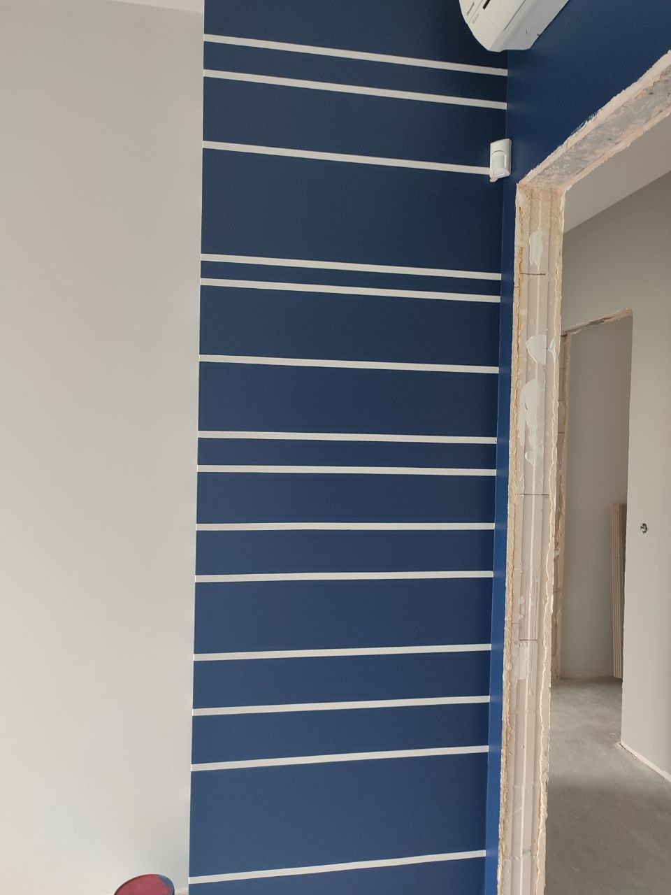 Granatowa ściana z białymi, poziomymi pasami, obok biała ściana i surowa framuga drzwi. Widoczny klimatyzator i czujnik.