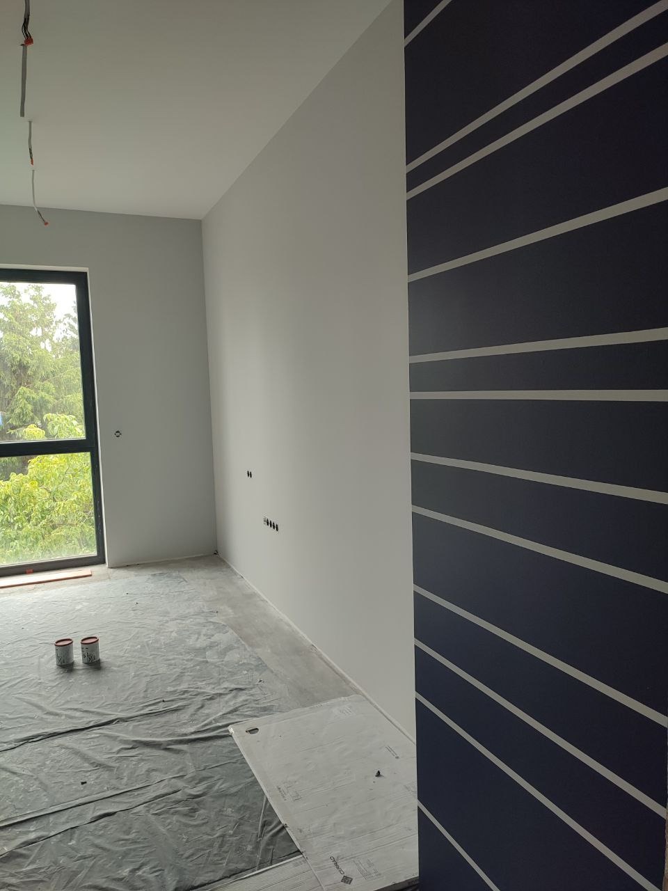 Wnętrze po malowaniu: ściany w odcieniach szarości i granatu z geometrycznym wzorem. Podłoga zabezpieczona folią, widoczne puszki farby.
