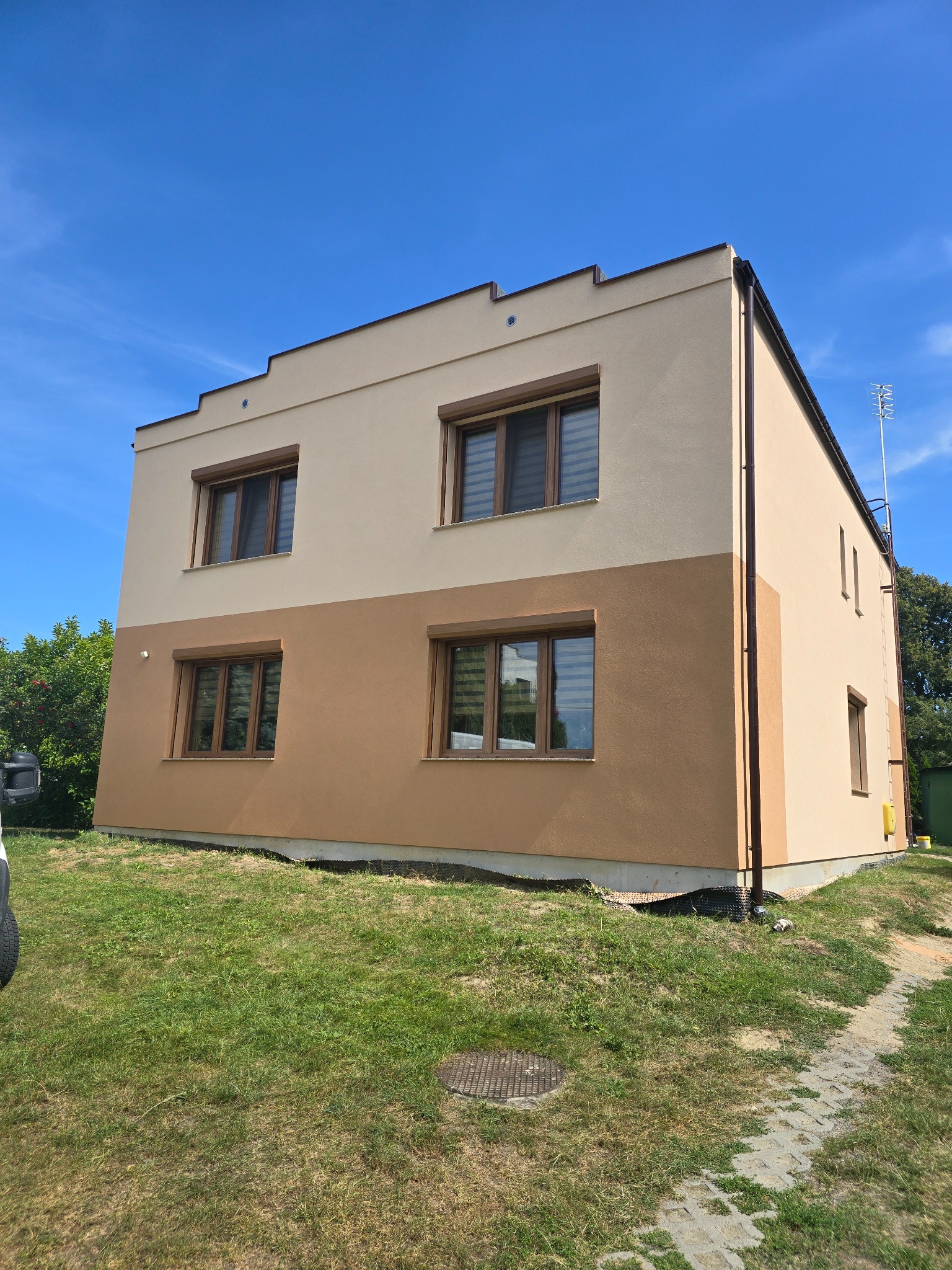 Dwukondygnacyjny dom z elewacją w dwóch odcieniach beżu, brązowe okna. Prace elewacyjne w Zaniemyślu. Widok z trawnika w słoneczny dzień.