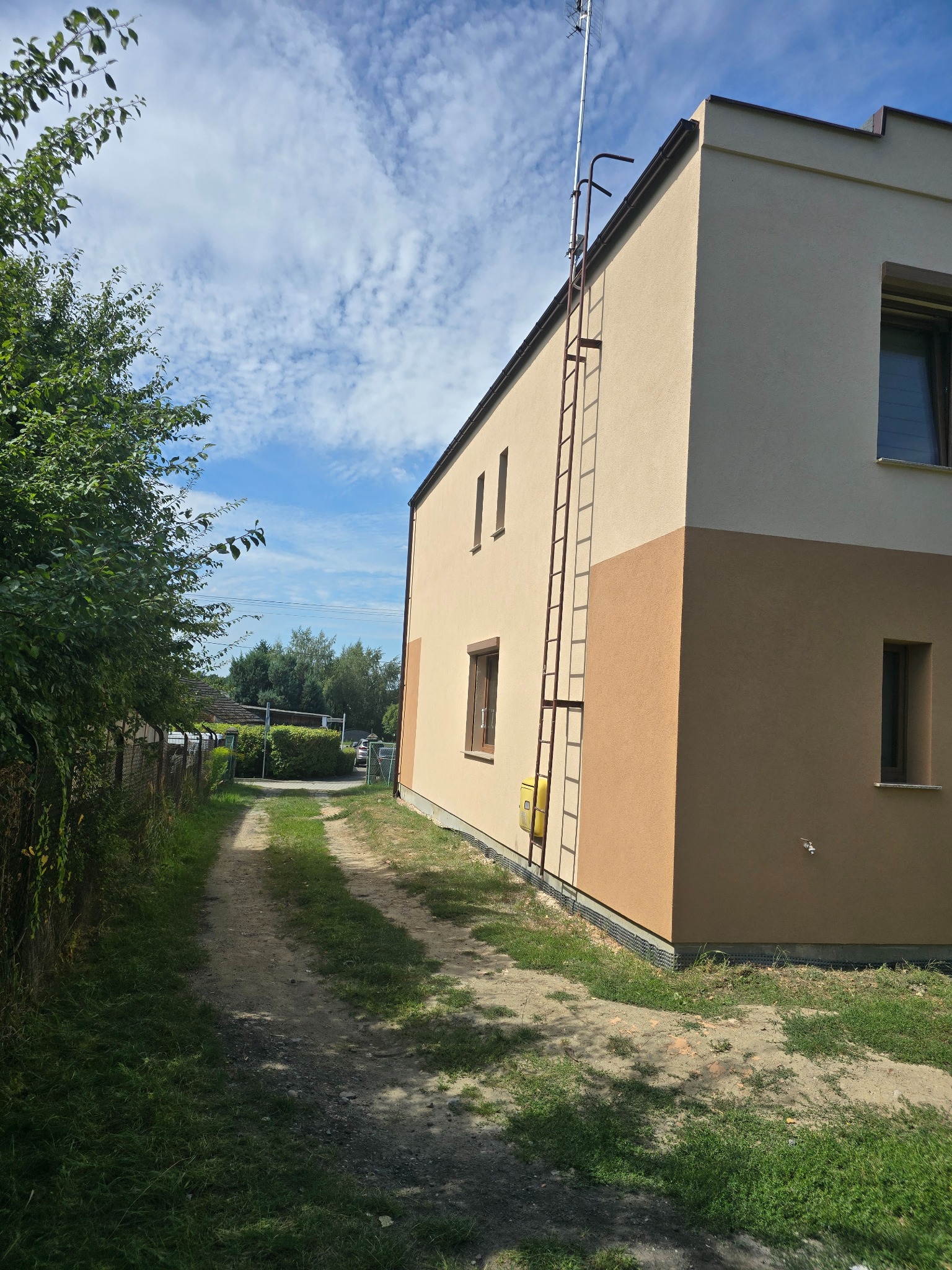 Dwukolorowa elewacja domu z brązowym dołem i beżową górą, z drabiną przymocowaną do ściany i anteną na dachu, widok z boku na tle zieleni i nieba.