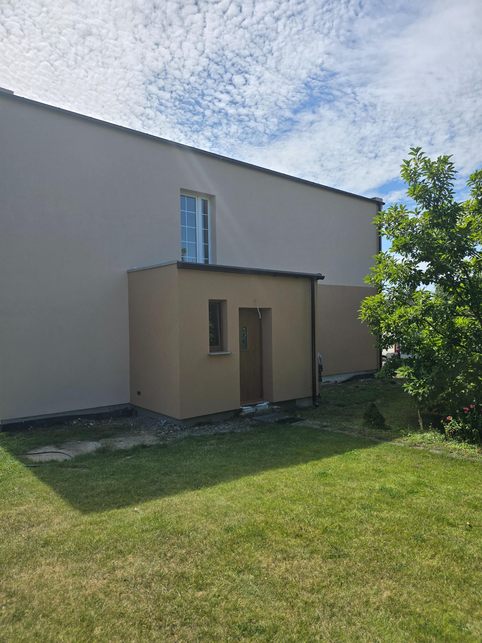 Otynkowana elewacja budynku z dobudówką w odcieniach beżu, widok z trawnika w słoneczny dzień. Białe okno na górze, brązowe drzwi dobudówki.