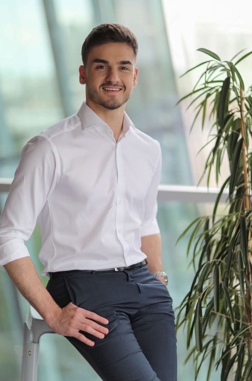 Uśmiechnięty mężczyzna w białej koszuli, oparty o krzesło na tle szklanej ściany i rośliny doniczkowej. Portret biznesowy w naturalnym oświetleniu.