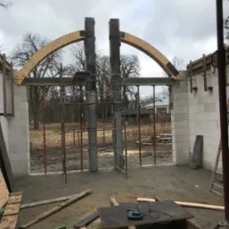 Wznoszenie łuku nad otworem w ścianie z betonu komórkowego. Konstrukcja wsparta na rusztowaniach. Na pierwszym planie narzędzia i deski.