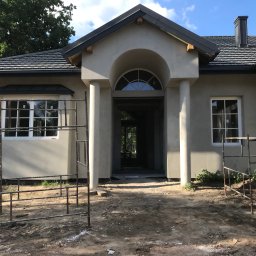 Usługi Ogólnobudowlane - Dom jednorodzinny w trakcie wykończenia elewacji; widoczne rusztowania, surowy tynk, czarna dachówka i okna z białymi ramami. Wejście z kolumnami i łukiem.