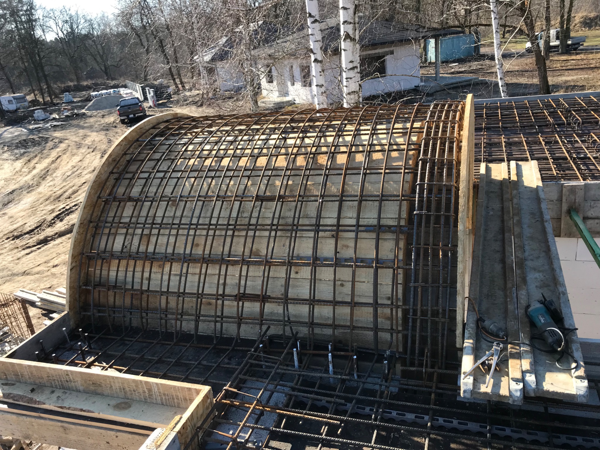 Zbrojenie łukowe z deskowaniem na budowie domu jednorodzinnego. Widoczne narzędzia i drewniane elementy konstrukcji. W tle drzewa i budynek.