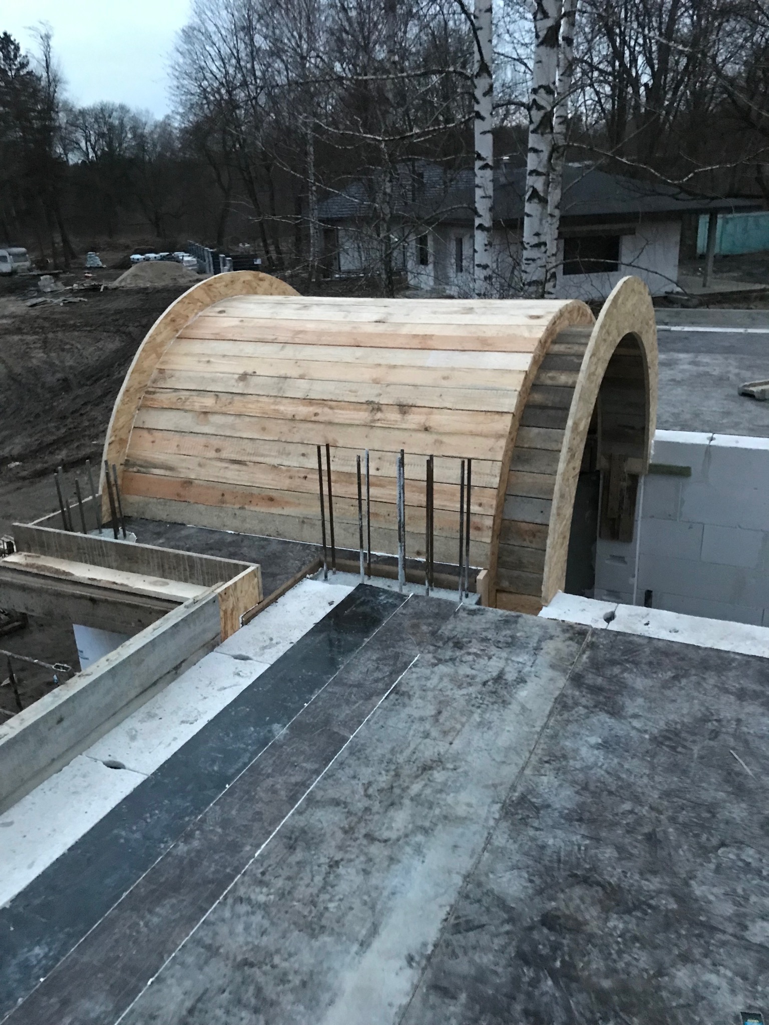 Konstrukcja łukowa z drewna i stali na betonowym fundamencie, etap budowy. W tle drzewa i budynek w surowym stanie.