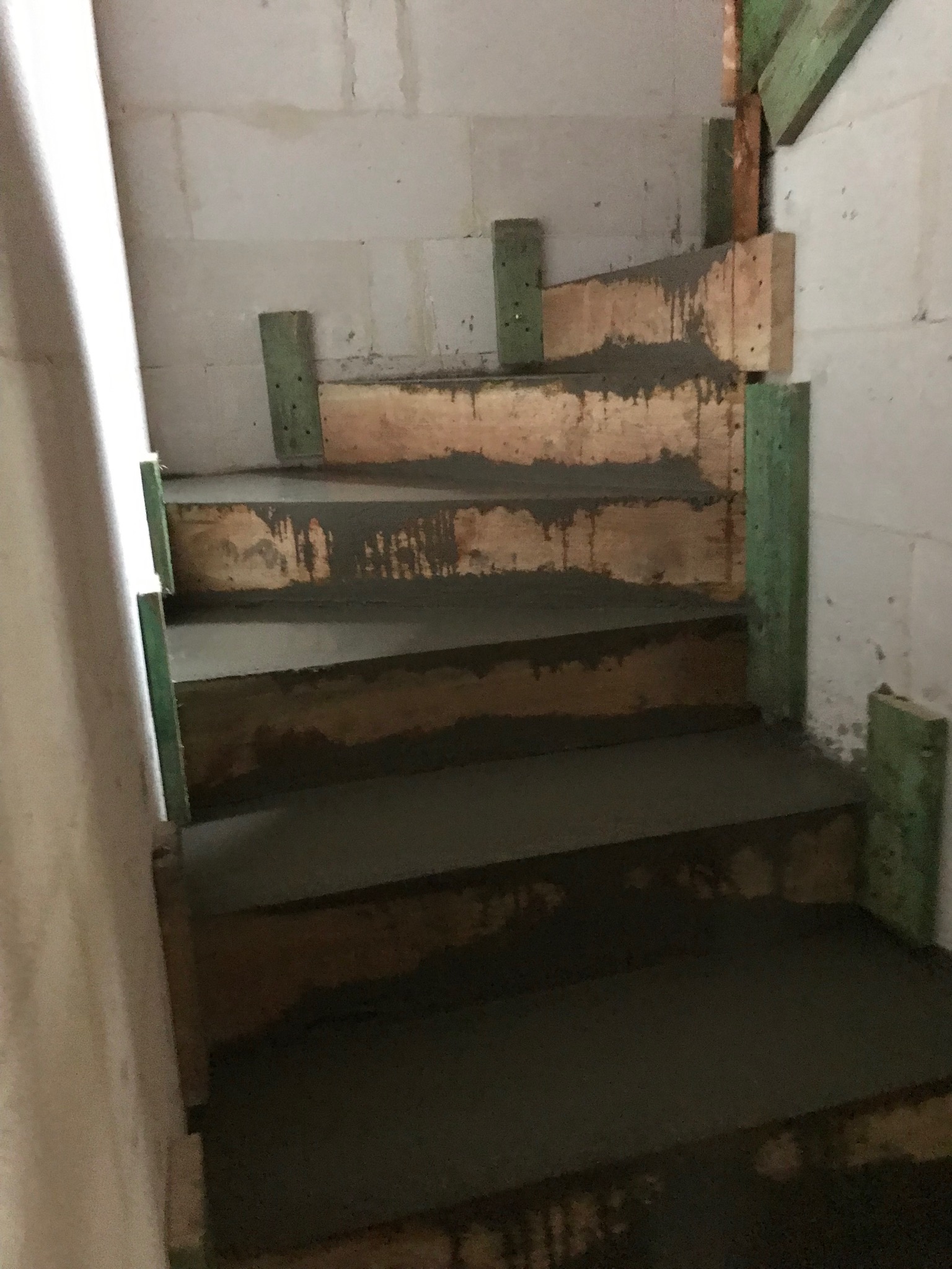 Betonowe schody w trakcie budowy, z drewnianym szalunkiem i zielonymi podporami, na tle ściany z bloczków betonowych.