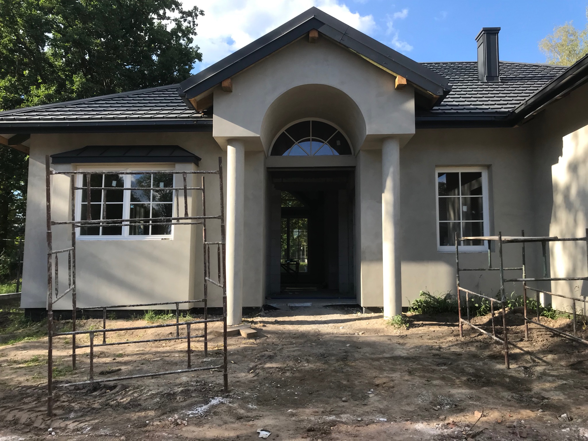 Dom jednorodzinny w trakcie wykończenia elewacji; widoczne rusztowania, surowy tynk, czarna dachówka i okna z białymi ramami. Wejście z kolumnami i łukiem.