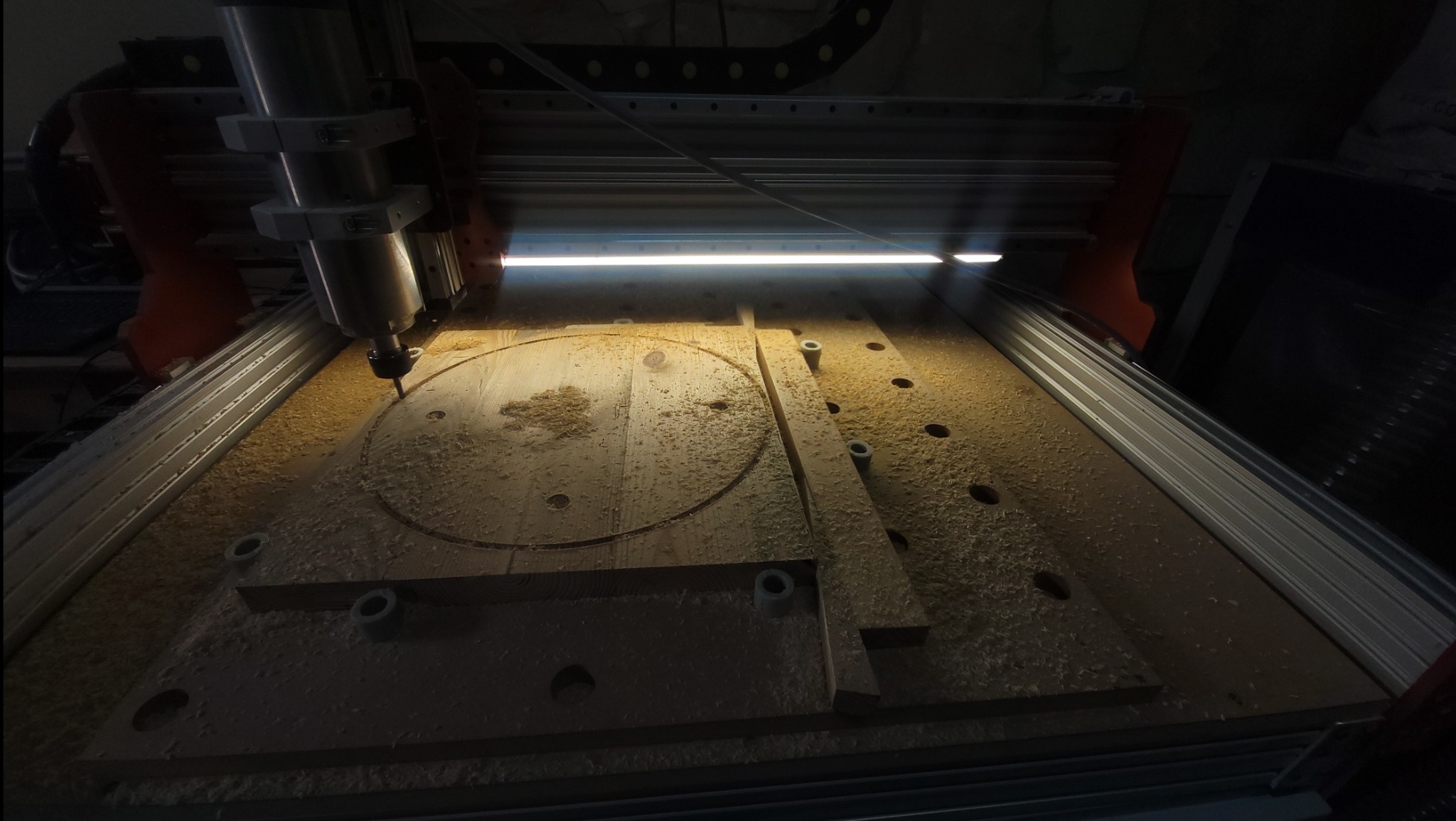 Frezarka CNC w trakcie pracy nad drewnianym elementem z wyfrezowanymi otworami i okręgiem, wióry na powierzchni, oświetlenie robocze.