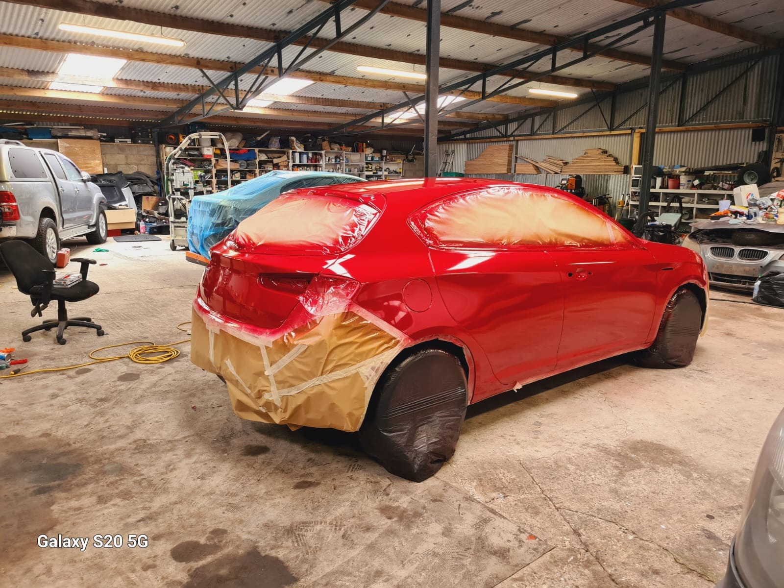 Czerwone auto w lakierni, zabezpieczone taśmą i folią przed malowaniem. Widoczne detale: świeży lakier, przygotowanie do dalszych prac.