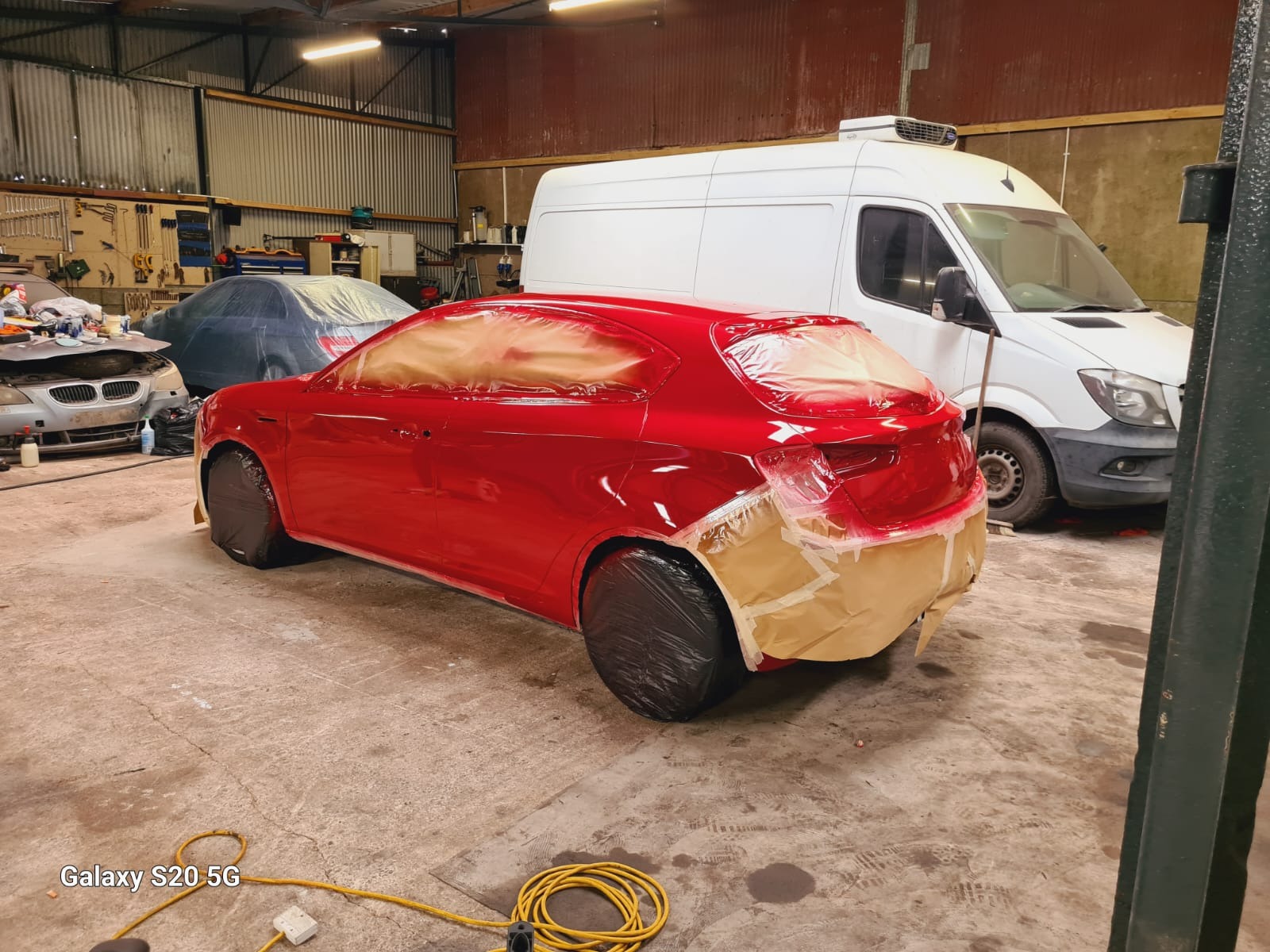 Czerwony samochód w trakcie lakierowania, zabezpieczony folią i taśmą, w warsztacie z widocznym wyposażeniem i białym busem w tle. Widoczny kabel zasilający na podłodze.