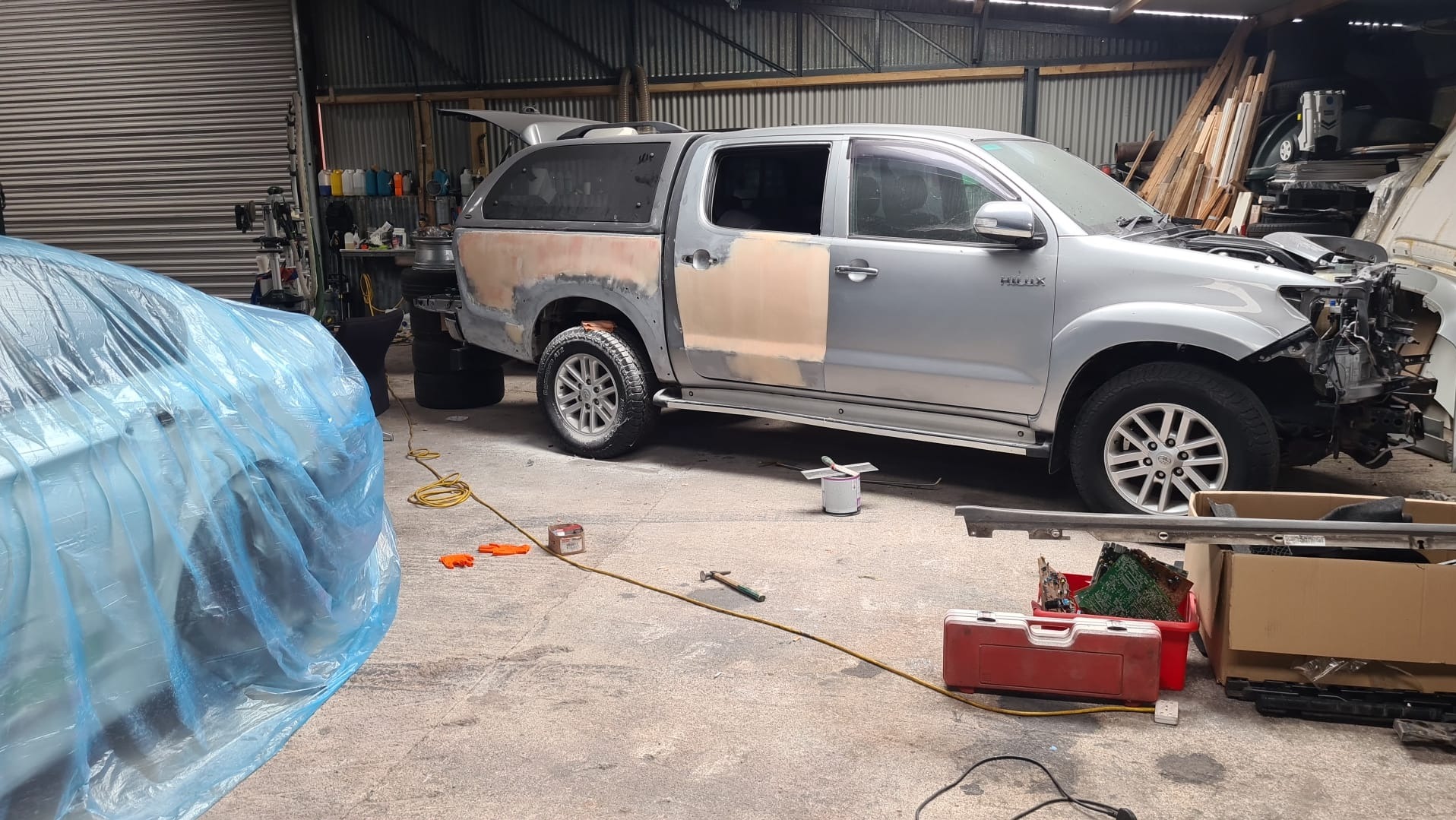 Srebrny pickup Hilux w warsztacie: przygotowania do lakierowania, zdemontowany przód, częściowo osłonięty folią samochód, narzędzia na podłodze.