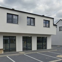 Nowoczesny budynek z szarymi oknami i przeszklonymi drzwiami wejściowymi. Część budynku w rusztowaniach. Parking z wyznaczonymi miejscami.