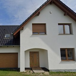 Biały dom jednorodzinny z brązową bramą garażową, oknami i roletą. Uwagę zwraca łukowe wejście z brązowymi drzwiami i rampą dla niepełnosprawnych.