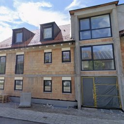 Nowy budynek w trakcie budowy z zamontowanymi oknami w czarnych ramach, widoczne rusztowania i materiały budowlane. Częściowo zamontowane rolety zewnętrzne.