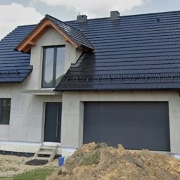 Marcin Mrowiec JACKY JO - Nowoczesny dom jednorodzinny w trakcie budowy, z szarą bramą garażową, oknami i drzwiami w tym samym odcieniu. Widoczne ocieplenie i przygotowanie pod elewację.