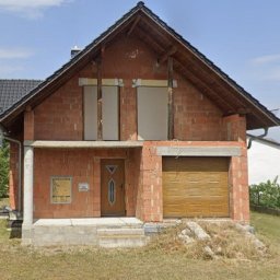 Marcin Mrowiec JACKY JO - Surowy stan domu jednorodzinnego z cegły, z zamontowaną bramą garażową i drzwiami wejściowymi. Okna częściowo zasłonięte, widoczne elementy konstrukcyjne.