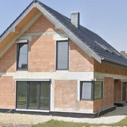 Dom w trakcie budowy z zamontowanymi oknami i drzwiami przesuwnymi. Widoczne bloczki ceramiczne, szara dachówka i częściowo zagospodarowany teren.