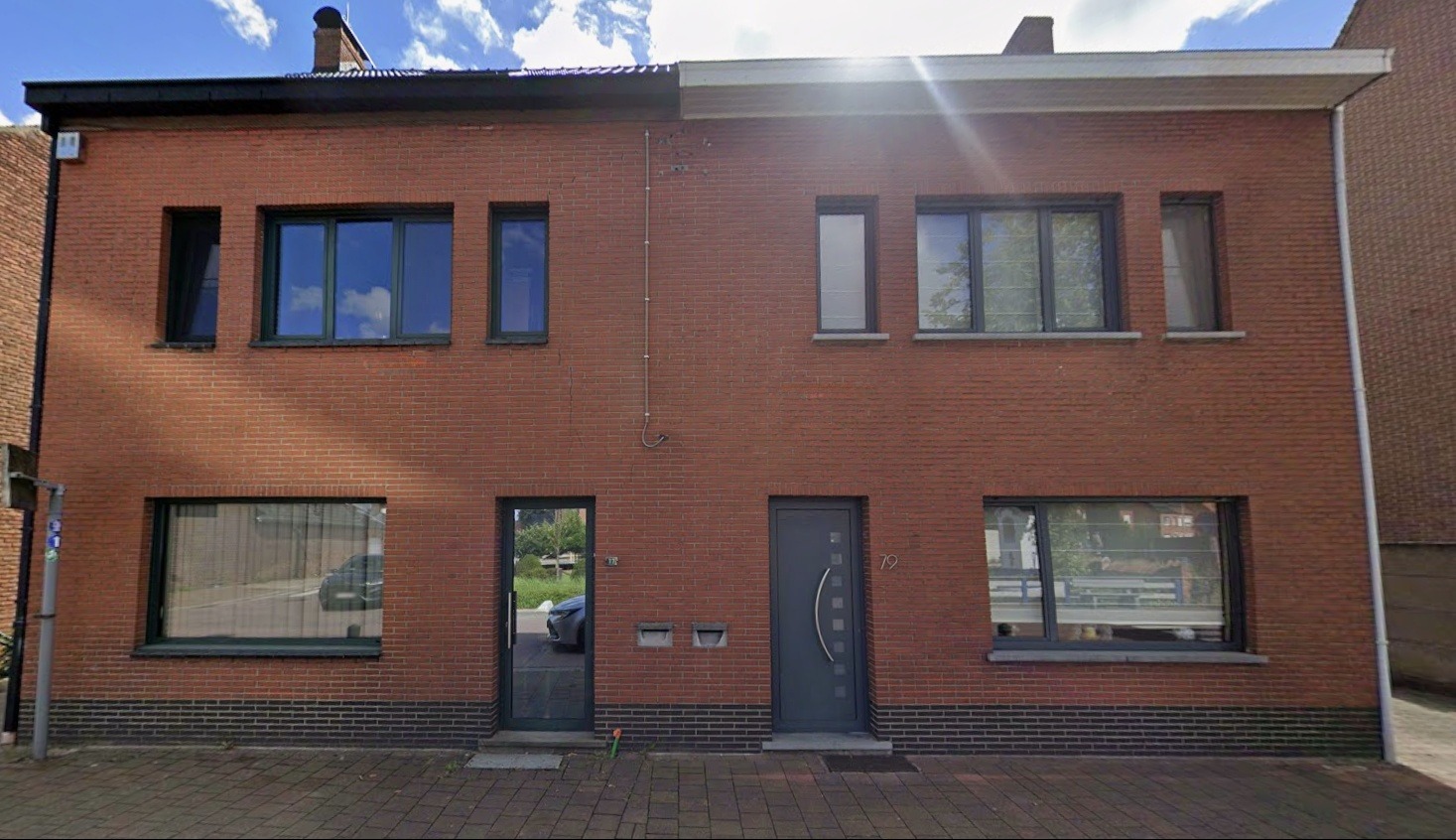 Ceglana fasada dwukondygnacyjnego domu z szarymi oknami i drzwiami. Po lewej stronie drzwi z widocznym odbiciem samochodu, po prawej drzwi z numerem 79.