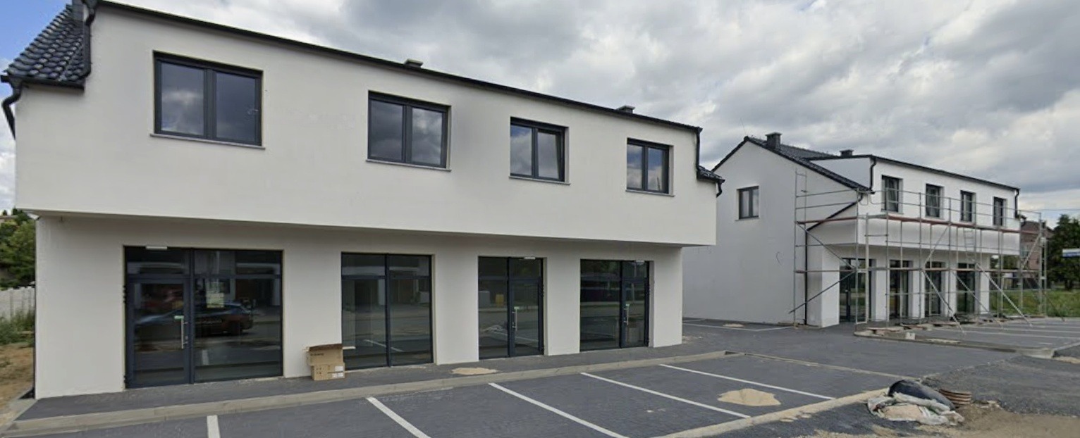 Nowoczesny budynek z szarymi oknami i przeszklonymi drzwiami wejściowymi. Część budynku w rusztowaniach. Parking z wyznaczonymi miejscami.