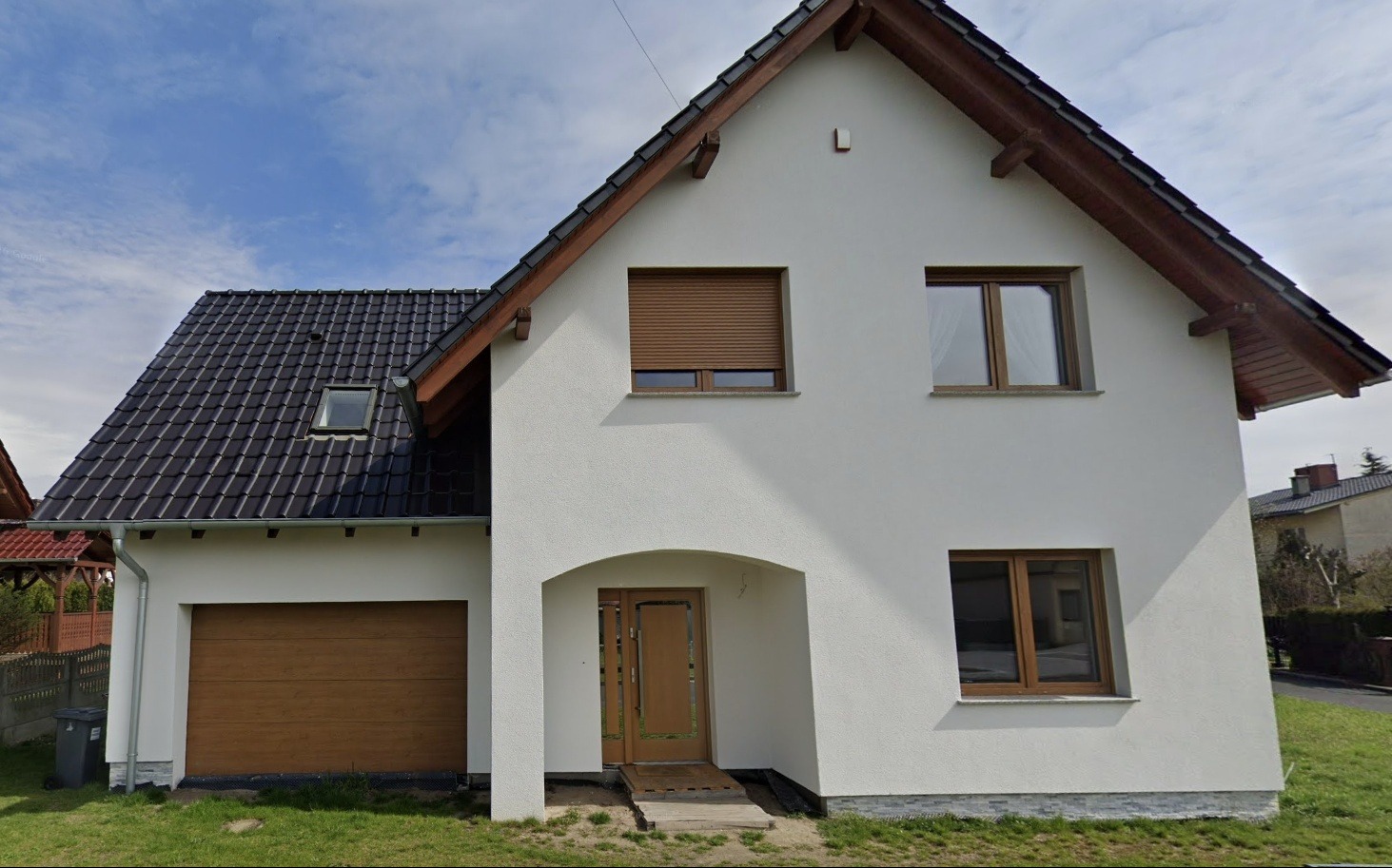 Biały dom jednorodzinny z brązową bramą garażową, oknami i roletą. Uwagę zwraca łukowe wejście z brązowymi drzwiami i rampą dla niepełnosprawnych.