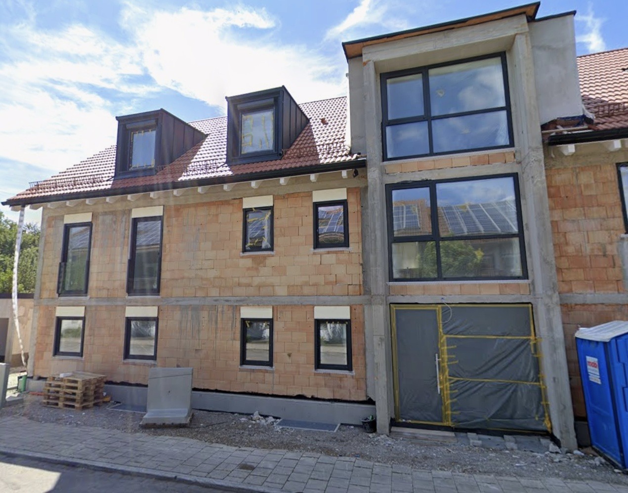 Nowy budynek w trakcie budowy z zamontowanymi oknami w czarnych ramach, widoczne rusztowania i materiały budowlane. Częściowo zamontowane rolety zewnętrzne.