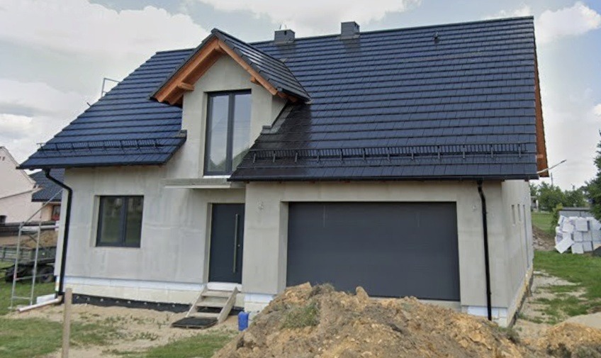 Nowoczesny dom jednorodzinny w trakcie budowy, z szarą bramą garażową, oknami i drzwiami w tym samym odcieniu. Widoczne ocieplenie i przygotowanie pod elewację.