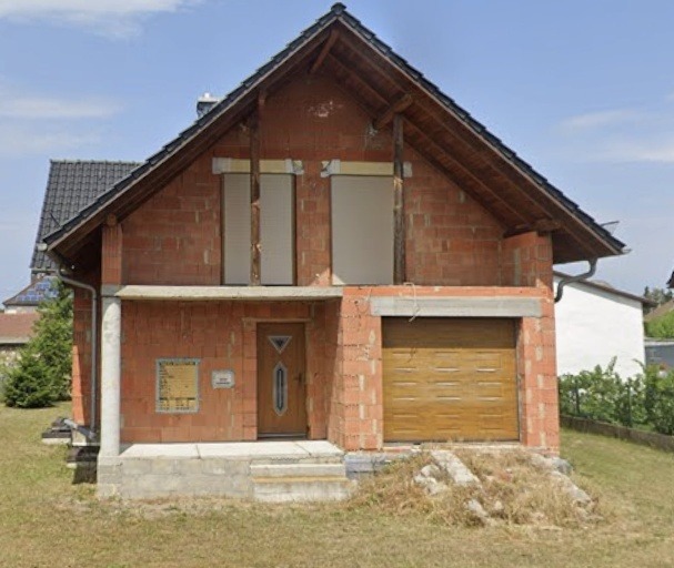 Surowy stan domu jednorodzinnego z cegły, z zamontowaną bramą garażową i drzwiami wejściowymi. Okna częściowo zasłonięte, widoczne elementy konstrukcyjne.